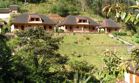 Drei charmante Hütten mit roten Dächern stehen auf einer sanften Wiese, umgeben von üppigem Grün und Bäumen.