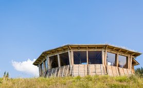 Ein modernes, rundes Holzgebäude mit großen Fenstern steht auf einer Wiese unter einem strahlend blauen Himmel.