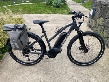 Ein modernes E-Bike mit einem grauen Rucksack steht auf einem gepflasterten Weg, umgeben von grünem Gras und Pflanzen.