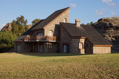 Ein modernes Steinhaus mit einem schrägen Dach steht in einer grünen Wiese, umgeben von sanften Hügeln und Bäumen.