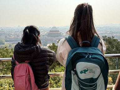 Zwei Personen stehen mit dem Rücken zur Kamera und blicken auf die Verbotene Stadt in Peking, umgeben von grünen Bäumen.