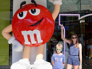 Ein großer, roter M&M's Charakter steht vor einem Geschäft, während ein Junge und ein Mädchen lächeln und posieren.