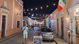 Eine belebte Straße in Valladolid, Mexiko, beleuchtet von Lichtern, mit einem Schild für Happy Hour und einer mexikanischen Flagge.