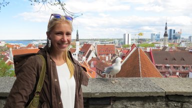 Eine lächelnde Frau steht an einer Mauer mit Blick auf die Altstadt von Tallinn und eine Möwe neben ihr.