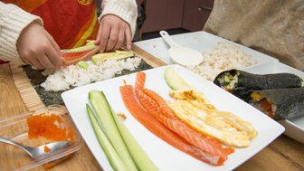Ein Kind rollt Sushi mit frischem Lachs, Avocado und Gurke auf einem Bambusmatte, während eine andere Person im Hintergrund zuschaut.