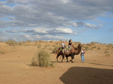 Eine Gruppe von Menschen reitet auf Kamelen durch eine trockene, sandige Landschaft unter einem bewölkten Himmel.