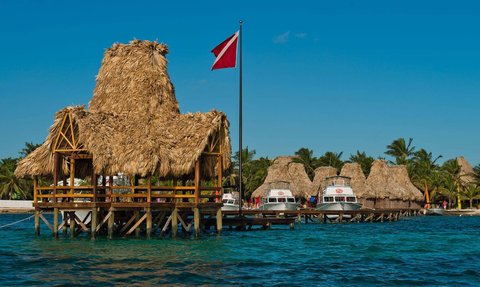 Ein malerisches Resort mit Strohdächern und Booten, umgeben von klarem Wasser und Palmen unter einem strahlend blauen Himmel.
