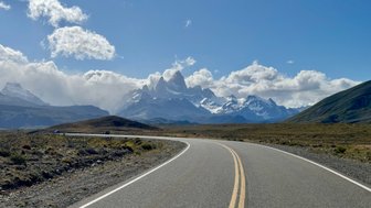 Eine kurvenreiche Straße führt durch eine atemberaubende Landschaft mit schneebedeckten Bergen im Hintergrund.