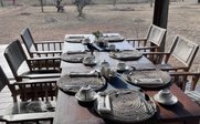 Ein rustikaler Tisch im Freien ist liebevoll gedeckt mit Geschirr und Besteck, umgeben von einer weiten, trockenen Landschaft.