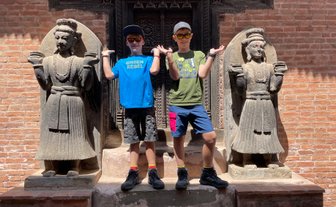 Zwei Jungen posieren fröhlich zwischen zwei beeindruckenden Steinfiguren in Bhaktapur, umgeben von rotem Ziegelmauerwerk.