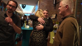 Eine Frau in einem glitzernden Kleid umarmt einen Mann herzlich, während andere Gäste im Hintergrund feiern.