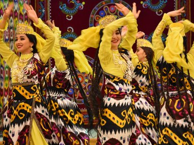 Fünf Tänzerinnen in bunten traditionellen Kleidern bewegen sich rhythmisch zur Musik, ihre Hände heben sich in die Luft.