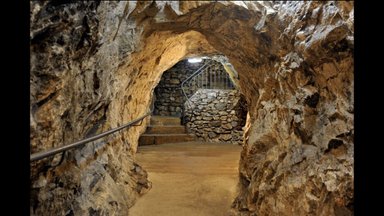 Ein schmaler, beleuchteter Tunnel mit steinernen Wänden und einer Treppe, die in die Tiefe führt, vermittelt ein geheimnisvolles Gefühl.