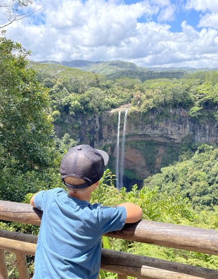 Ein Junge steht an einem Geländer und blickt auf den beeindruckenden Chamarel-Wasserfall inmitten üppiger Natur.