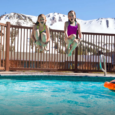 Zwei fröhliche Mädchen springen gleichzeitig in einen klaren Pool, umgeben von schneebedeckten Bergen im Hintergrund.