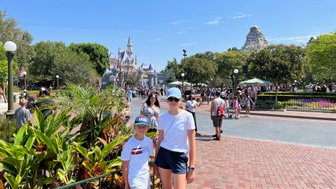 Zwei Kinder stehen vor dem Disneyland-Park, umgeben von bunten Pflanzen und fröhlichen Besuchern an einem sonnigen Tag.