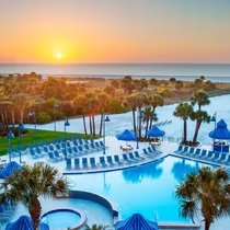 Ein atemberaubender Sonnenuntergang über einem Pool, umgeben von Palmen und blauen Liegen, schafft eine entspannende Atmosphäre.