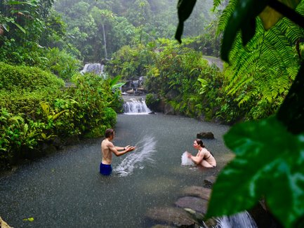 Inmitten eines üppigen Dschungels spielen zwei Personen im Wasser, während strömender Regen um sie herum fällt.