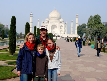 Eine Familie posiert fröhlich vor dem Taj Mahal, umgeben von grünen Bäumen und einem klaren Himmel.