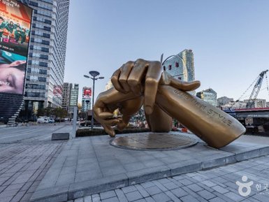 Eine große, goldene Statue einer Hand, die ein Schild mit der Aufschrift 'Gangnam Style' hält, steht in einer urbanen Umgebung.
