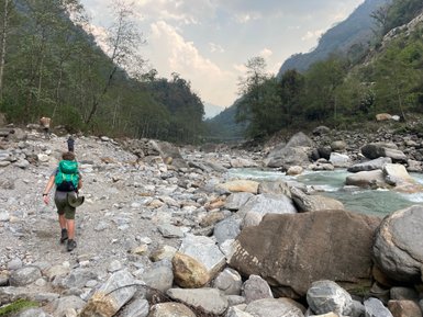 Ein Wanderer mit einem grünen Rucksack geht über einen steinigen Weg entlang eines Flusses in den Bergen Nepals.