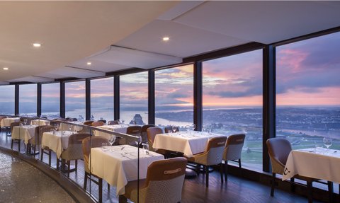 Ein elegantes Restaurant mit Blick auf die Stadt, umgeben von großen Fenstern, die einen atemberaubenden Sonnenuntergang zeigen.