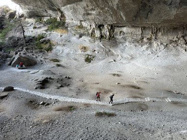 Zwei Wanderer gehen auf einem schmalen Pfad unter einer großen Felswand, während im Hintergrund eine Gruppe sitzt.