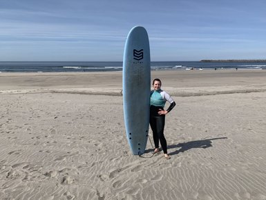 Eine Surferin steht am Strand mit einem blauen Surfbrett, umgeben von sanften Wellen und einem klaren Himmel.