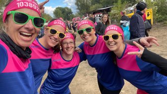 Fünf Frauen posieren fröhlich für ein Selfie, tragen bunte Sportkleidung und Sonnenbrillen, umgeben von einer lebhaften Veranstaltung.