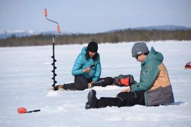 Zwei Personen sitzen auf dem Eis, während eine Frau mit einem Angelgerät in der Hand konzentriert arbeitet.