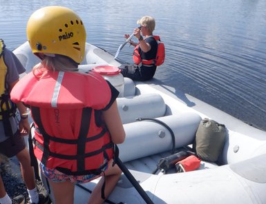 Eine Gruppe von Personen bereitet sich auf eine Raftingtour auf einem ruhigen Gewässer mit einem Schlauchboot vor.
