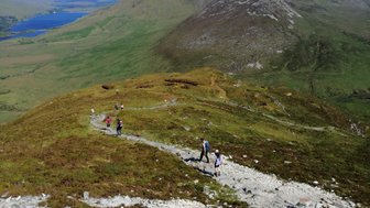 Eine Gruppe von Wanderern erkundet einen malerischen Bergpfad mit grünen Wiesen und einem klaren blauen See im Hintergrund.