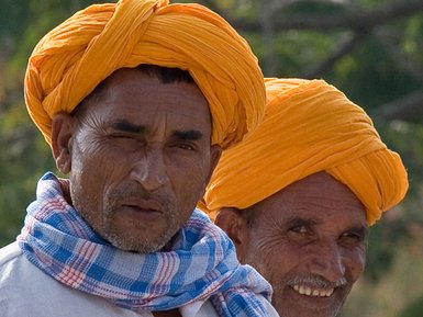 Zwei Männer mit leuchtend orangefarbenen Turbanen und Schals stehen im Freien, einer lächelt, während der andere ernst schaut.