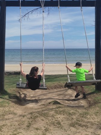 Eine Frau und ein Kind schwingen glücklich an einer Schaukel am Strand, umgeben von sanften Wellen und klarem Himmel.