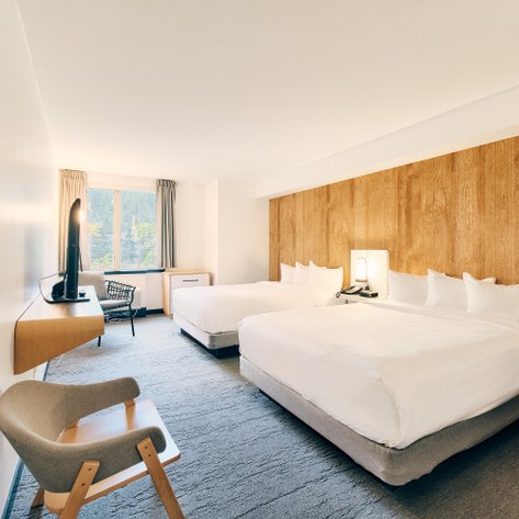 Ein modernes Hotelzimmer mit zwei großen Betten, einem Schreibtisch und einem Fenster, das auf eine grüne Landschaft blickt.