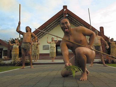 Ein Māori-Krieger zeigt seine Zunge, während er eine Pflanze auf dem Boden hält, umgeben von Tänzern in traditioneller Kleidung.