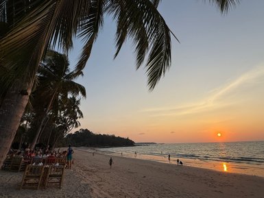 Ein malerischer Strand bei Sonnenuntergang, umgeben von Palmen und Menschen, die am Wasser spazieren gehen.