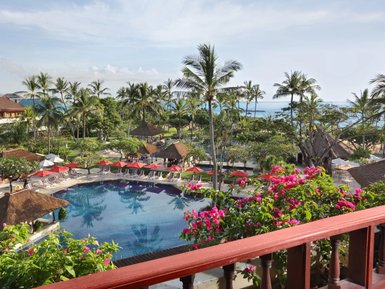 Ein Blick auf den Pool des Nusa Dua Beach Hotels, umgeben von Palmen und bunten Blumen, mit Sonnenliegen und roten Sonnenschirmen.