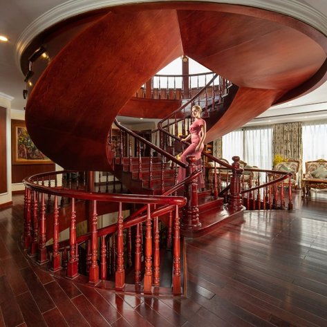 Eine elegante Frau in einem roten Kleid steht auf einer geschwungenen Treppe aus Holz in einem luxuriösen Raum.