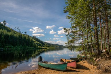 Ein ruhiger See umgeben von dichten Wäldern, mit bunten Kanus am Ufer, die auf das glitzernde Wasser blicken.