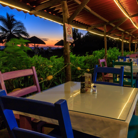 Ein einladendes Restaurant am Strand mit bunten Stühlen und Tischen, umgeben von üppigem Grün und einem Sonnenuntergang im Hintergrund.