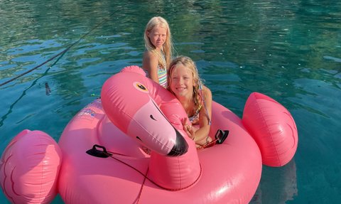 Zwei fröhliche Mädchen sitzen auf einem großen, pinken Flamingo-Schwimmring im klaren Wasser einer Bucht.