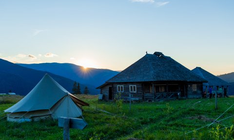 Ein Zelt steht auf einer Wiese, während die Sonne hinter den Bergen aufgeht und ein Holzhaus im Hintergrund sichtbar ist.
