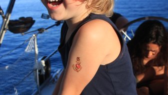 Ein fröhlicher Junge mit blonden Haaren lacht, während er auf einem Segelboot steht, mit einem Anker-Tattoo auf dem Arm.