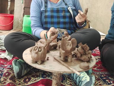 Eine lächelnde Frau mit grauen Haaren sitzt auf einem Teppich und formt Tonfiguren auf einem Tisch.