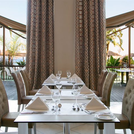 Elegante Restaurant mit schön gedeckten Tischen, Blick auf den Garten und das Meer durch große Fenster.