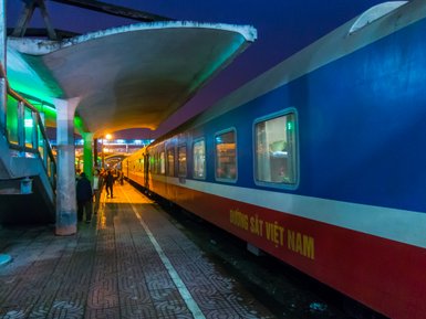 Ein Nachtzug steht an einem beleuchteten Bahnhof, während Passagiere auf dem Bahnsteig warten und sich unterhalten.