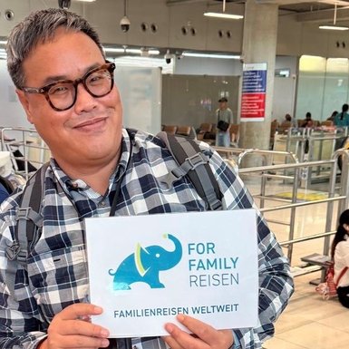 Ein lächelnder Reiseleiter hält ein Schild mit der Aufschrift 'FOR FAMILY REISEN' am Flughafen in einer belebten Umgebung.