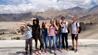 Eine Gruppe von Teenagern posiert fröhlich vor einer beeindruckenden Berglandschaft mit Wolken am Himmel.