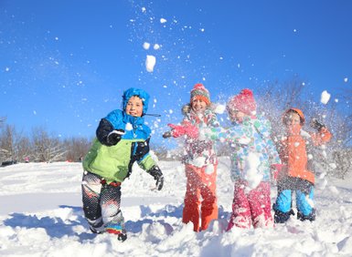 Vier fröhliche Kinder spielen im Schnee, werfen Schneebälle und lachen unter einem strahlend blauen Himmel.